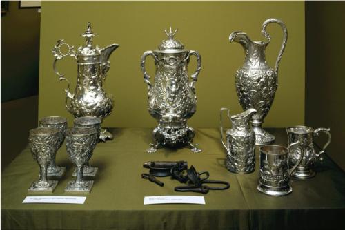 Figure 2.1 Display of silver tea service and associated silver objects on a table covered with green felt, arranged next to a set of metal shackles used to imprison enslaved Black persons